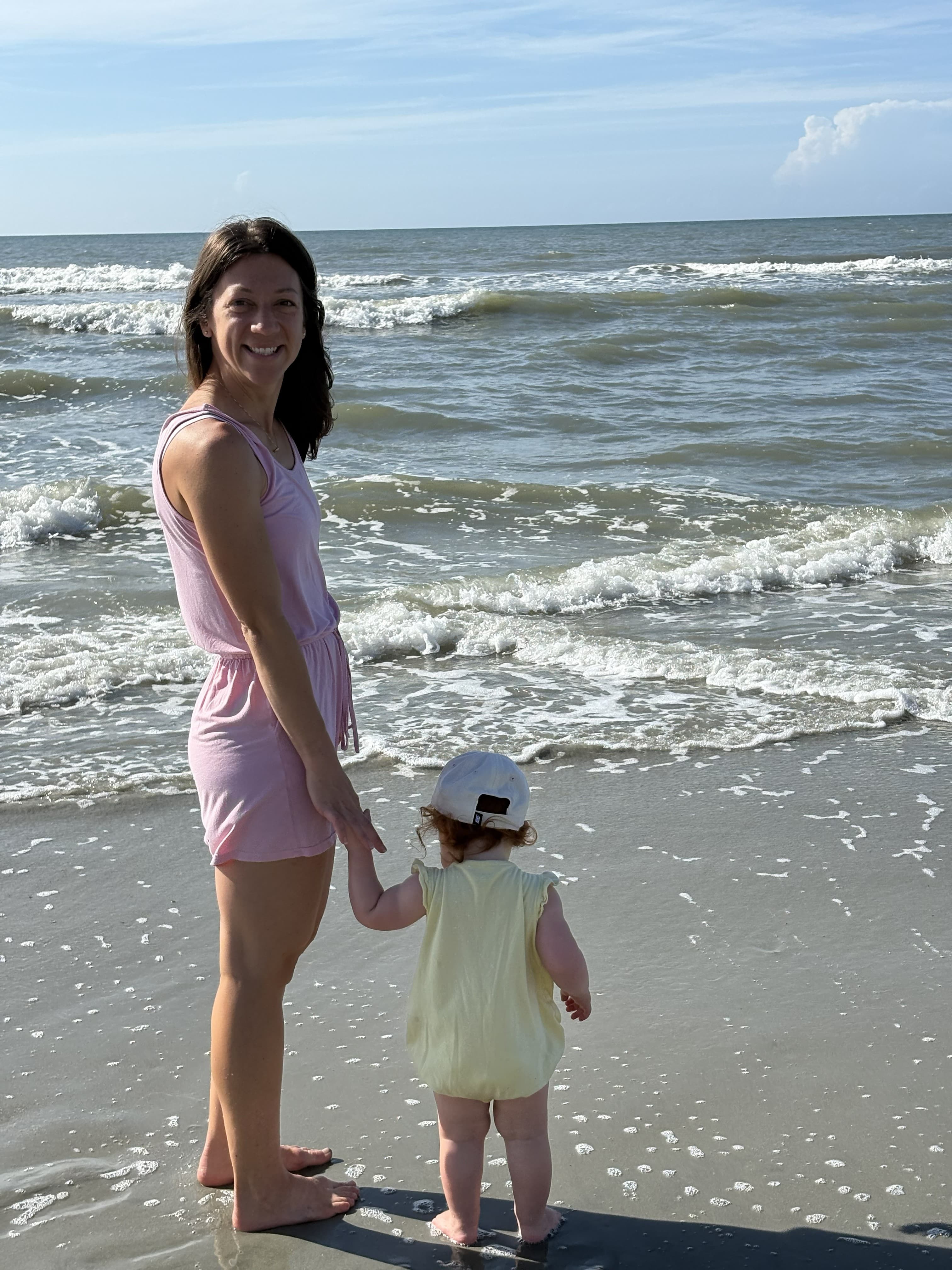 Smiling woman in a pink romper holds a toddler's hand at the ocean's edge.