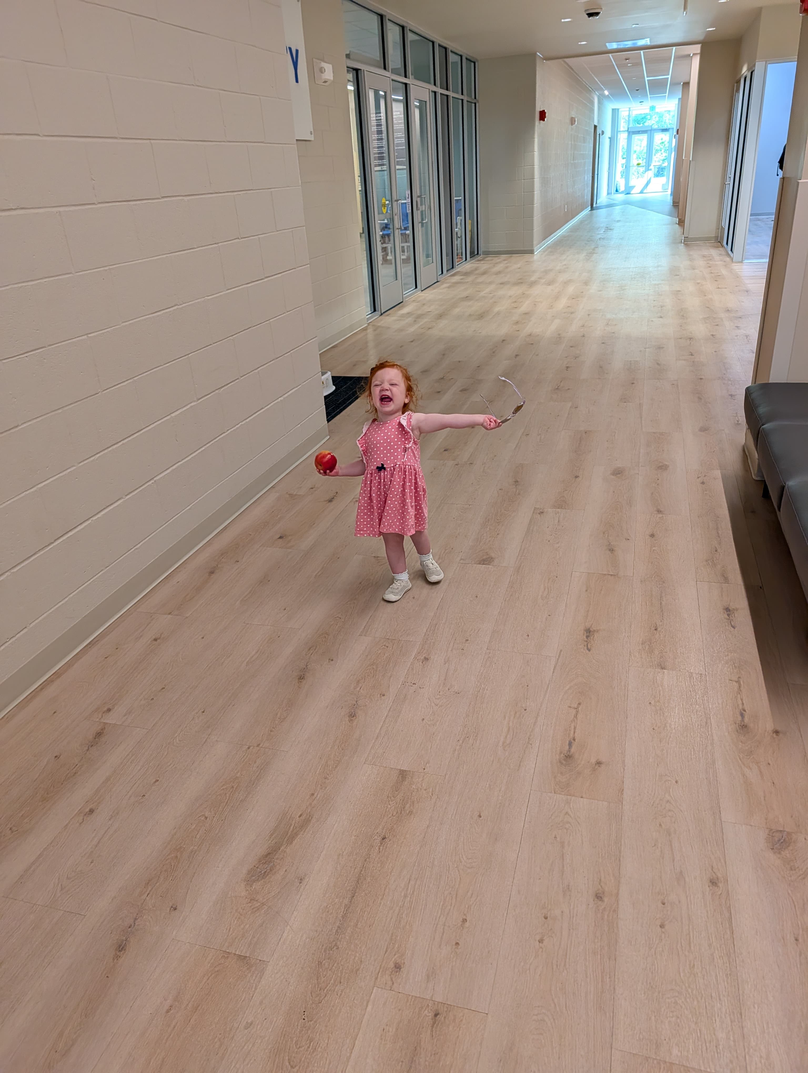 Red-haired toddler in a pink dress shouting, holding an apple and glasses in a hallway.