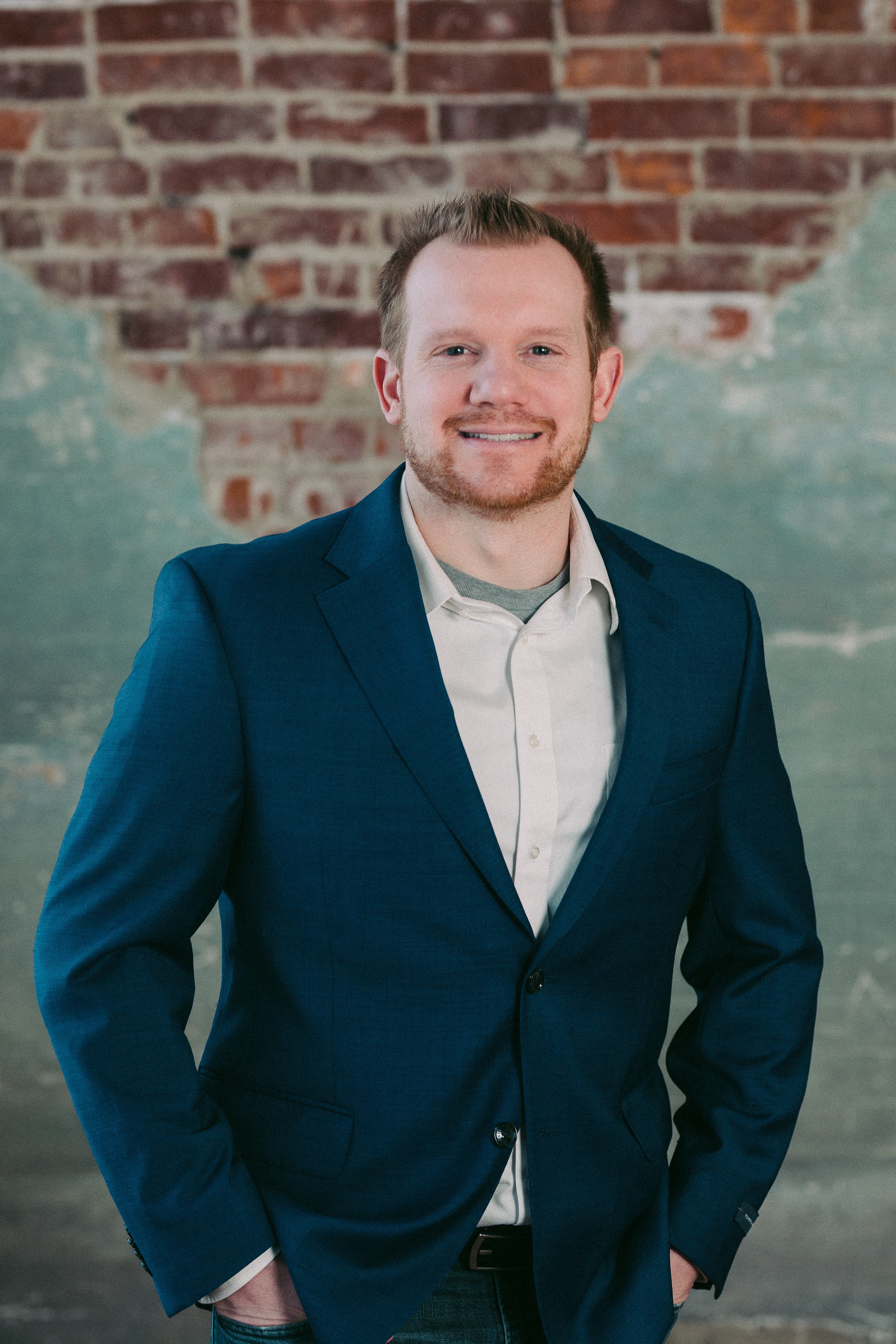 Smiling man with a beard in a blue blazer against a weathered brick wall.